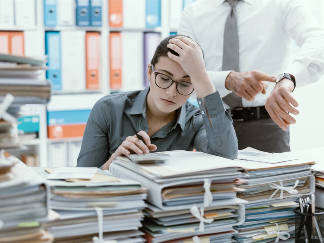 An exasperated worker at their desk, while their micromanaging boss stands behind indicating the time on a watch.