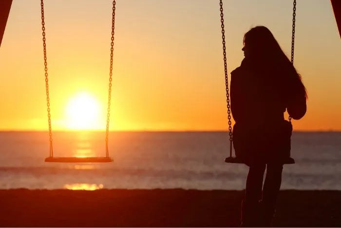 Silhouette of two swings at sunset, one empty and one with a person sitting in it.